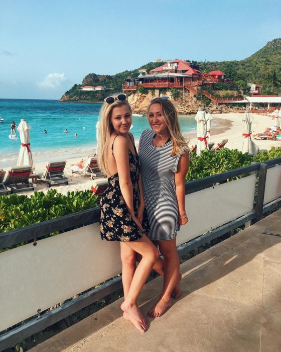 Madre e hija rubias posando descalzas en el malecón frente a la playa, ambas con vestidos playeros y lentes de sol