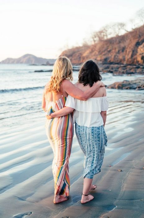 Madre de cabello castaño e hija rubia caminando descalzas en la arena en la playa mientras se abrazan