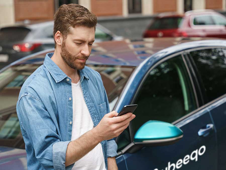 A bearded man is looking at his mobile phone, behind him a blue car with the Ubeeqo logo