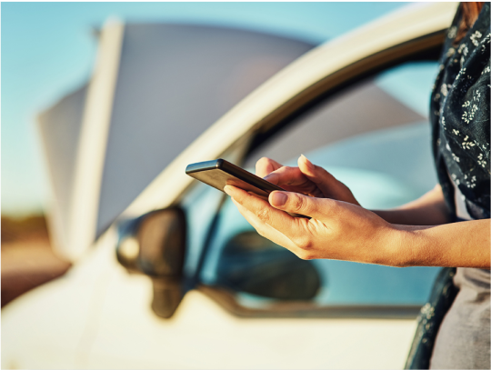 A woman's hand with blue fingernails holding a mobile phone, with a grey car in the background