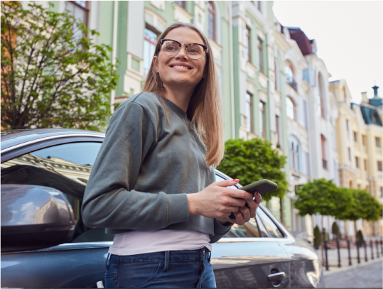 Un home amb barba mira el seu mòbil, darrere d'ell hi ha un cotxe blau amb el logo d'Europcar On Demand