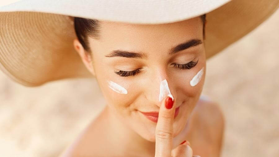 woman wearing a large stylish hat putting on sunscreen on her face