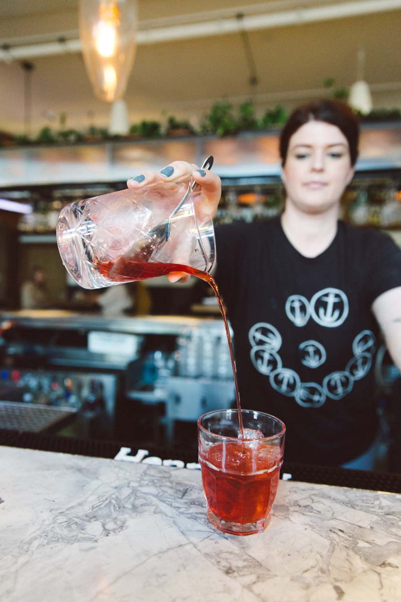woman behind a marble bar top pouring a negroni from a mixing glass into a rocks glass