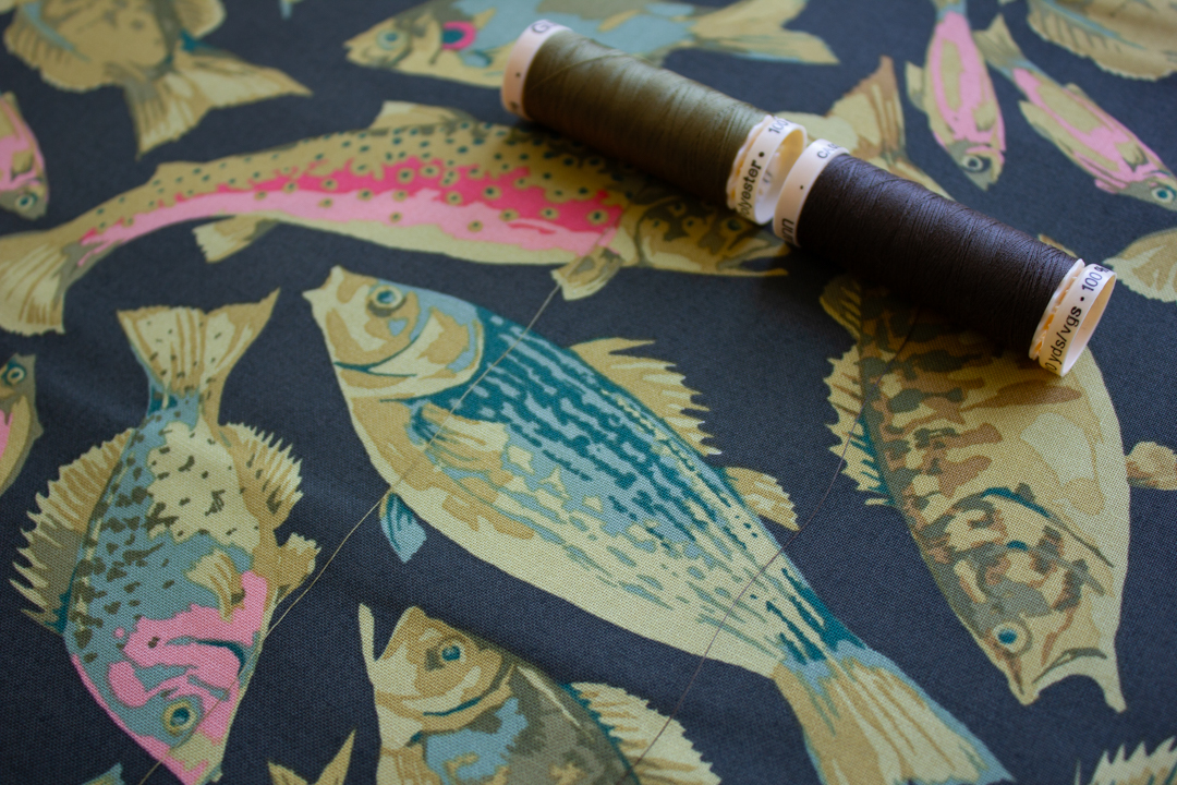 Two spools of thread, an olive green and charcoal gray, unwound a bit to show that the colors match the background fabric, which is a large-scale illustrated print of multicolored fish. 