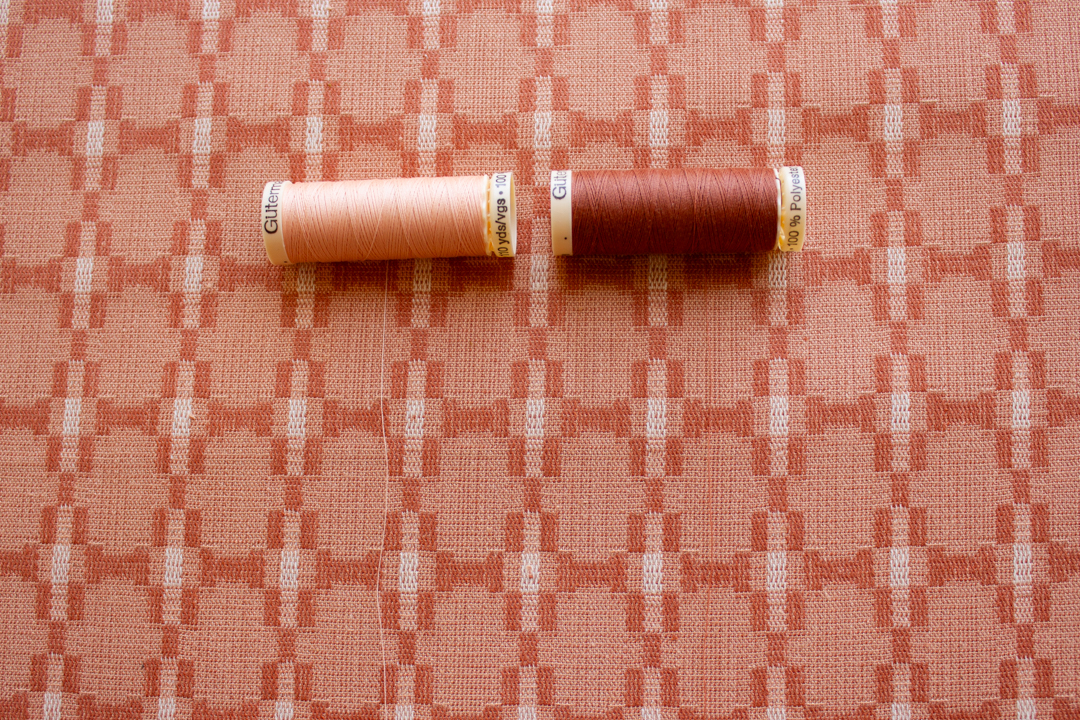 Photo of two spools of thread, a lighter peach and a darker peach, partially unrolled on a yarn-dyed background fabric in various shades of peach, to show that the thread colors match the fabric. 