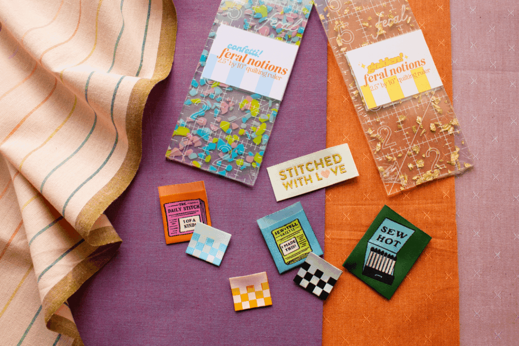 Colorful fabric swatches and sewing patches arranged on a vibrant background, featuring a 2.5 by 10-inch quilting ruler from Feral Notions, along with playful labels like "Stitched with Love" and various themed patches ideal for sewing enthusiasts.