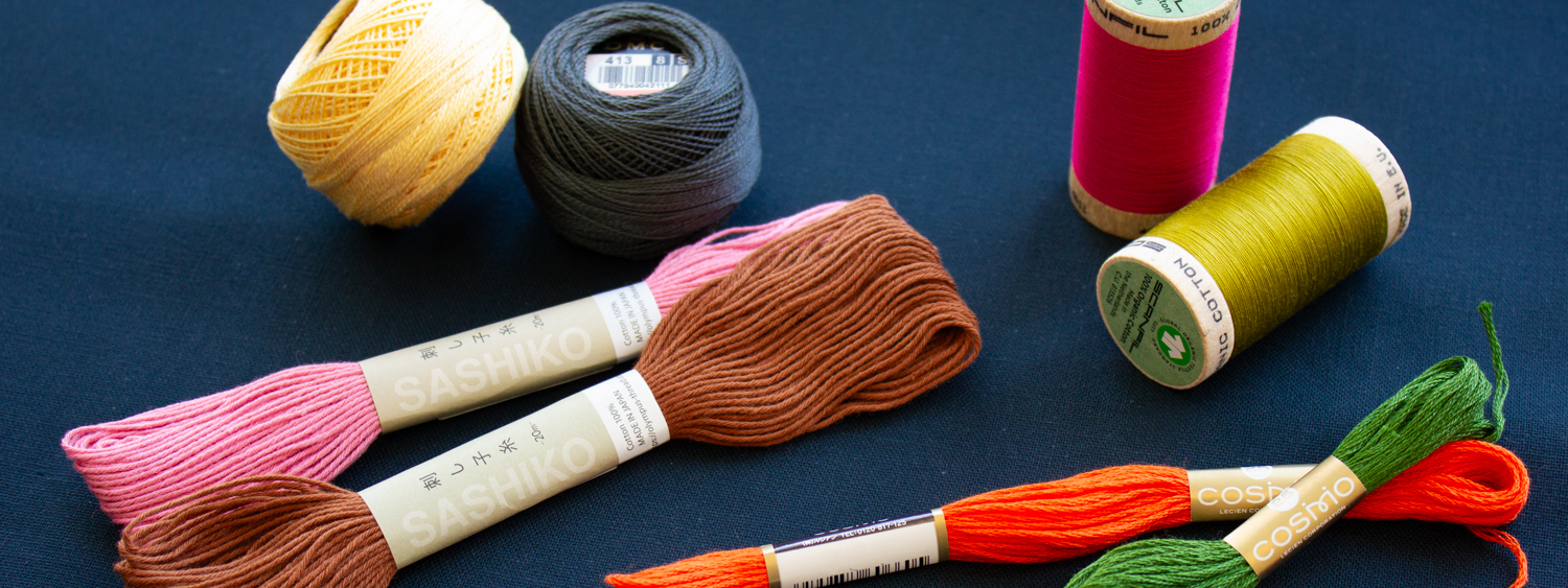 Colorful embroidery threads and yarn spools arranged on a dark blue background, featuring Sashiko and Cosmo brands in various shades including pink, brown, yellow, green, and orange, ideal for sewing and crafting projects.