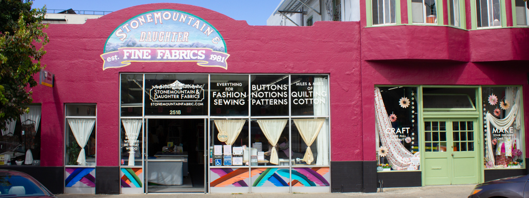 Exterior view of Stone Mountain & Daughter Fabrics, a vibrant fabric store established in 1981, featuring a colorful storefront with large windows displaying various fabric options, sewing notions, and craft supplies. The store offers a wide selection for fashion sewing, quilting, and crafting enthusiasts.