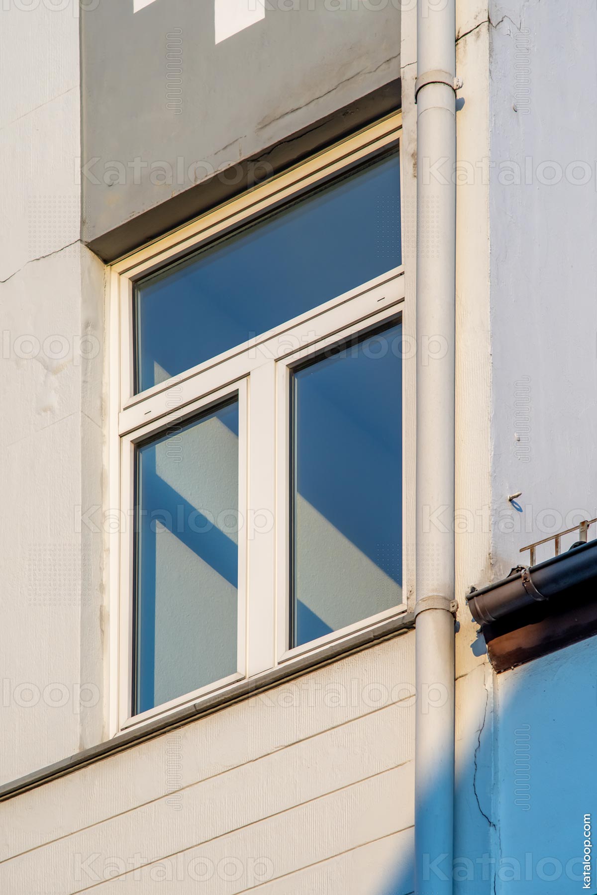 This image captures the play of light and shadow on the facade of a building, creating an abstract geometric pattern. A window with white frames is partially visible, contrasting with the textured blue wall. The scene is a blend of architectural elements and the natural interplay of sunlight.