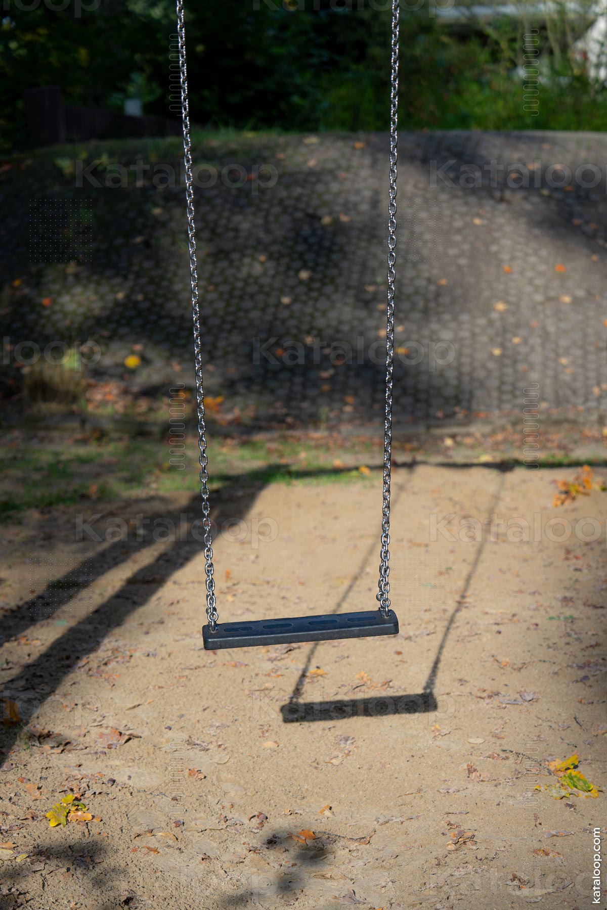 This image captures a solitary swing hanging from metal chains on a playground. The ground is covered with sand and scattered autumn leaves, suggesting a quiet, possibly chilly day. The swing casts a long shadow, indicating either early morning or late afternoon light.
