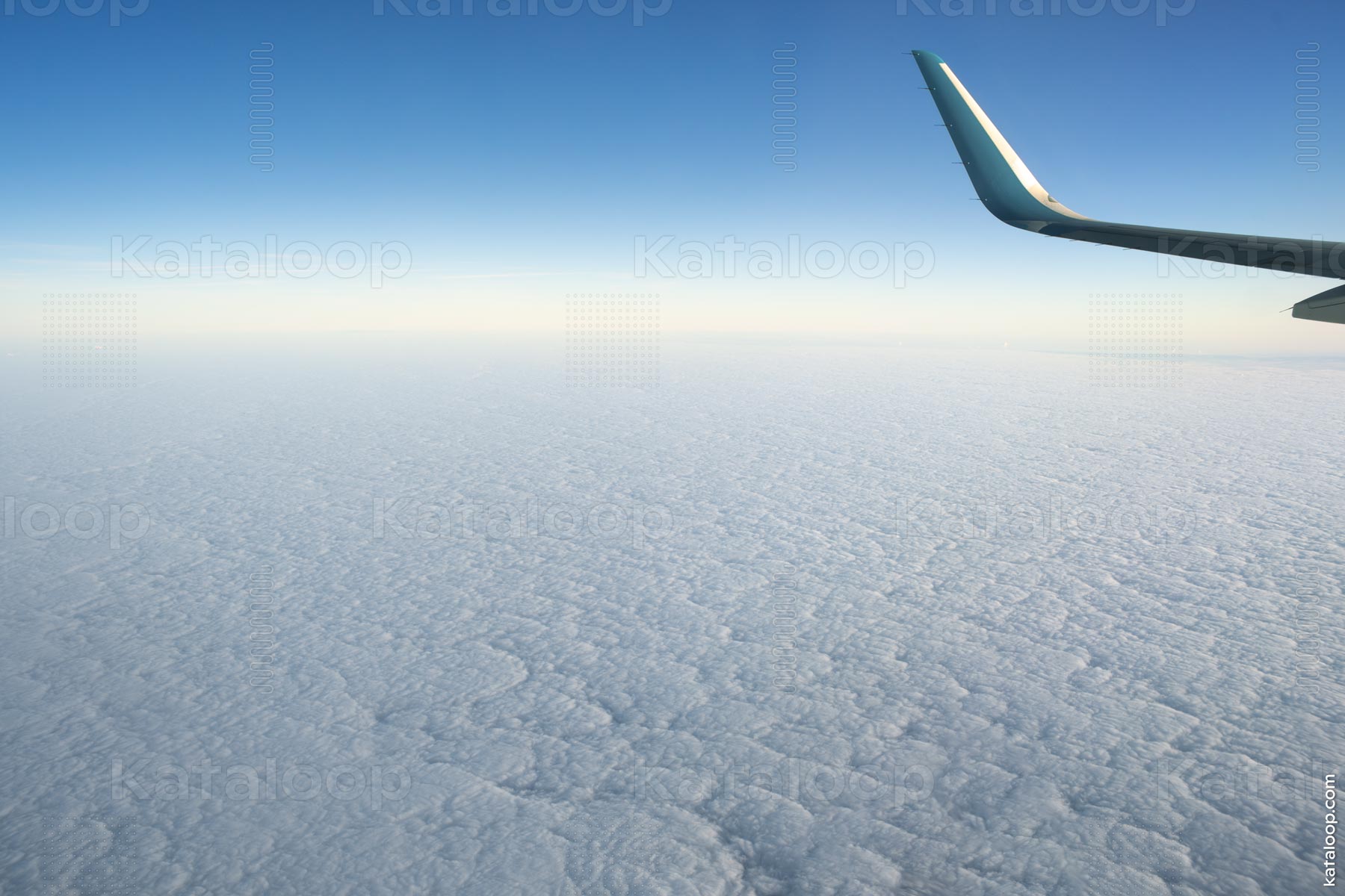 This image captures the serene beauty of a cloudscape as seen from the window of an airplane. The vast expanse of clouds blankets the earth, creating a soft, textured surface that stretches to the horizon. The airplane's wing tip enters the frame from the right, adding a sense of scale and travel to the scene.