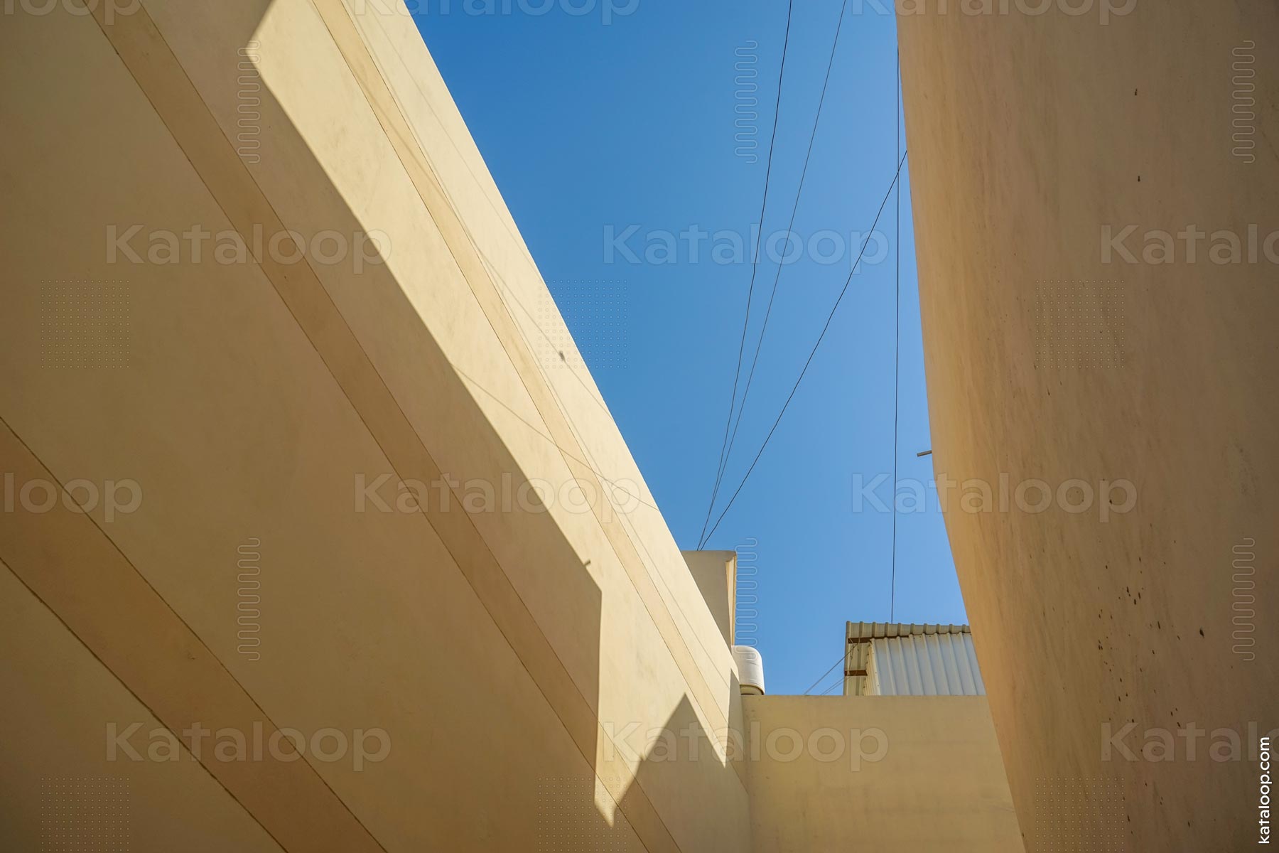 This image captures the interplay of light and shadow on modern architectural structures in Manama, Bahrain. The beige walls and clear blue sky create a striking contrast, highlighting the geometric shapes. Overhead wires add an urban element to the scene.