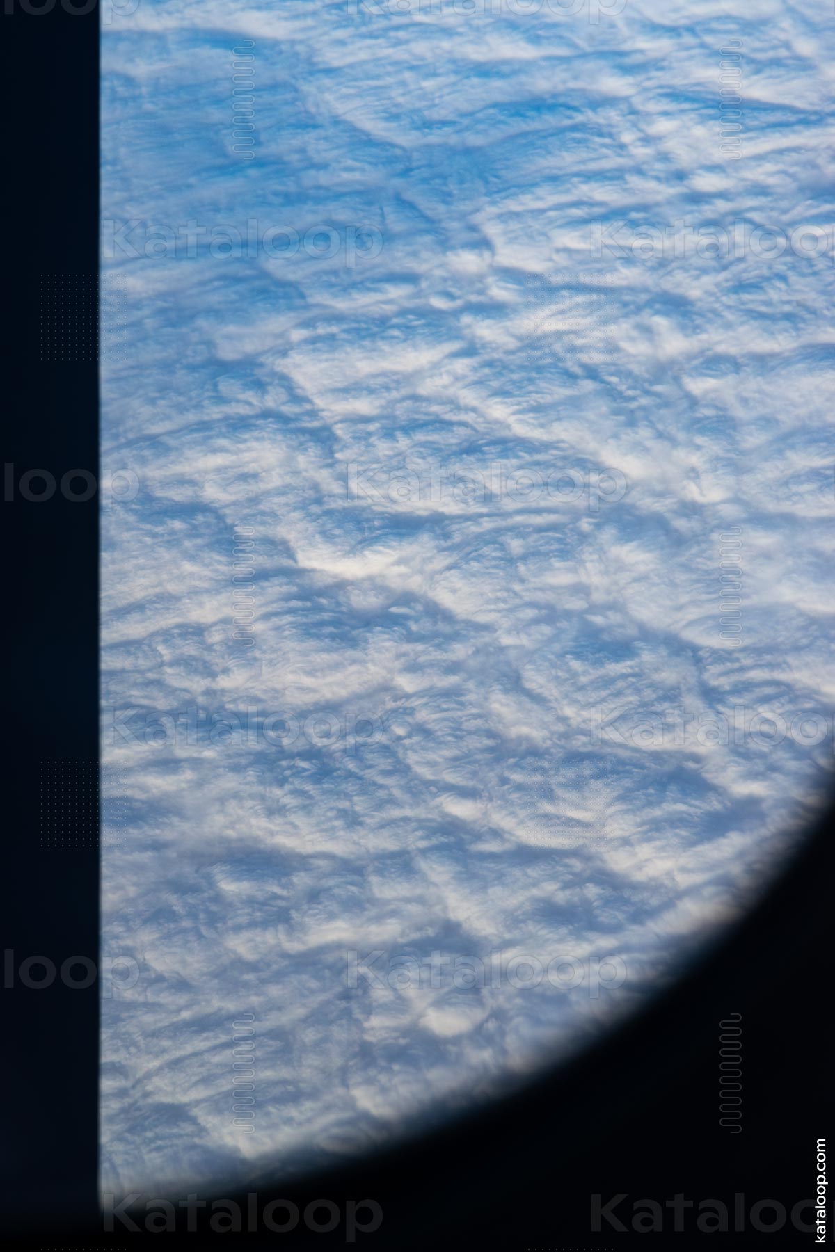 This image captures the serene beauty of a cloudscape as seen from the perspective of an airplane passenger. The frame of the airplane window is visible, providing a unique vantage point of the fluffy, textured clouds spread out below. The varying shades of blue and white create a tranquil and expansive atmosphere.
