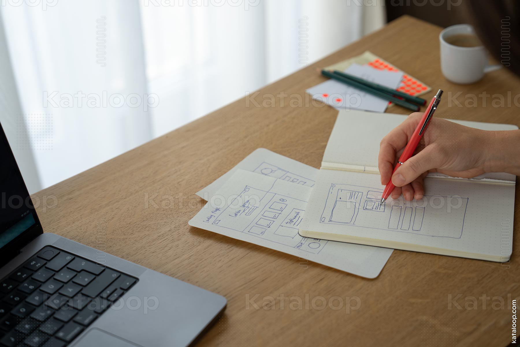 This image captures the process of designing a website layout with a close-up view of a hand-drawn wireframe in a notebook. The person is sketching different elements of a webpage, indicating a creative brainstorming session. A laptop is partially visible, suggesting the use of technology in the design process.