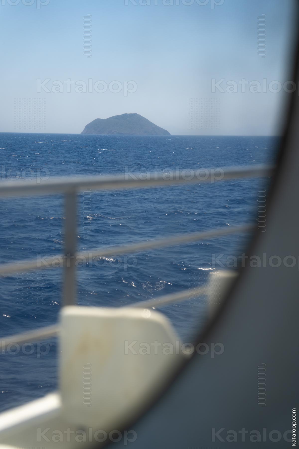 The image captures a scenic view of Strongyli Island as seen from a ferry window. The deep blue sea stretches towards the island, with the ferry railing visible in the foreground. This serene scene is perfect for travel and nature enthusiasts.