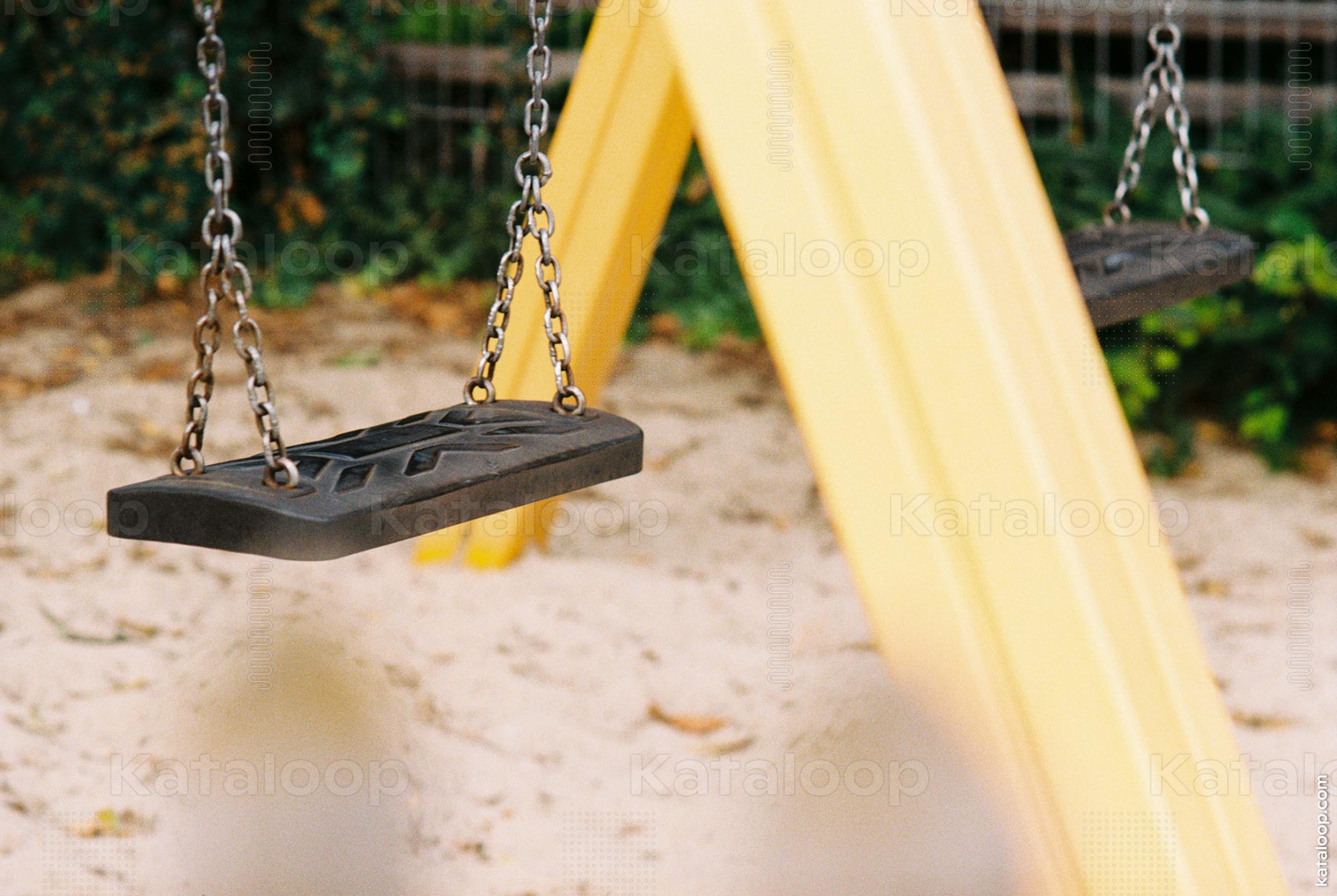 The image shows an empty swing set in a playground. The swing is made of black rubber and is suspended by metal chains. The background features a sandy ground and a yellow support structure. This scene captures a quiet moment in a children's play area, ideal for themes of childhood, leisure, and outdoor activities.