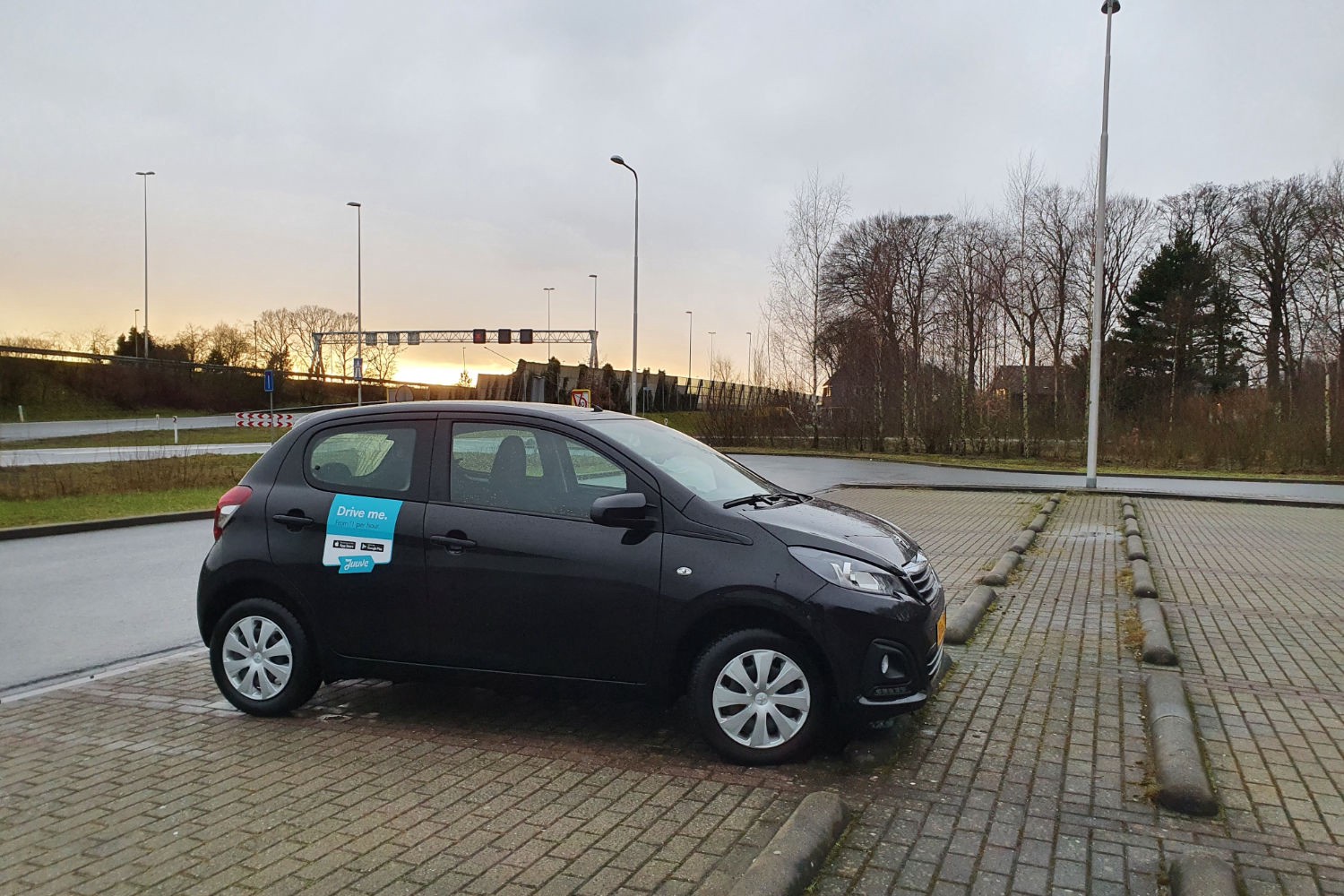 Photo of a Peugeot 108 parked in a parking lot next to a motorway. It has a sticker on it that says Juuve, the name of the rental company.