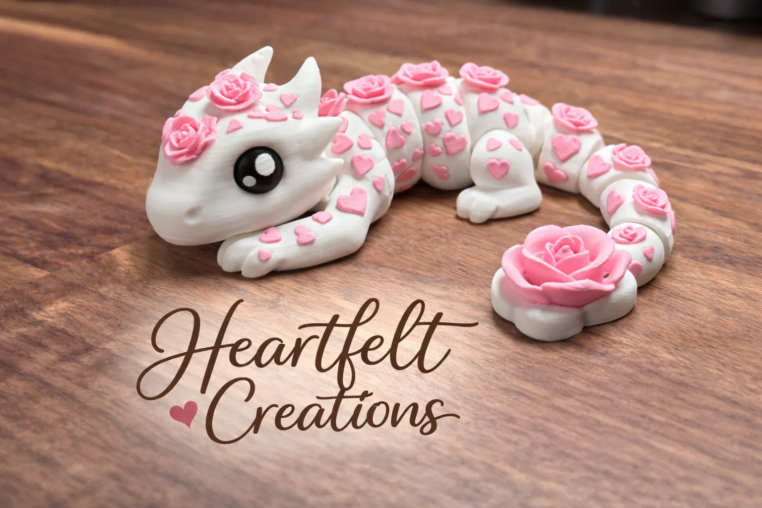 A cute, white 3D printed dragon figurine covered in small pink hearts and topped with tiny pink roses along its back and tail base. It has large black eyes and rests on a dark wood surface next to the Heartfelt Creations logo.