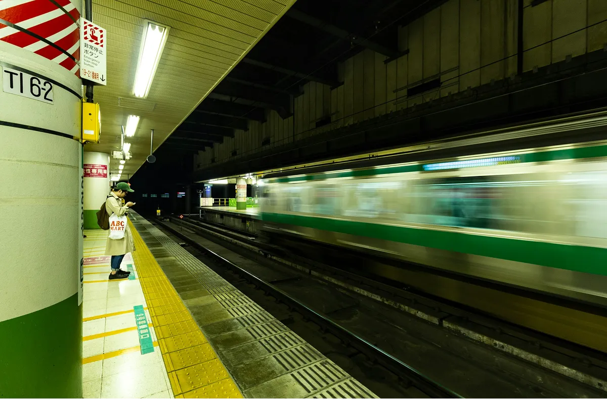 駅のホームで電車を待つ人の姿。通過中の緑色の電車がモーションブラーで流れるように写っている。黄色い点字ブロックが敷かれたホーム。