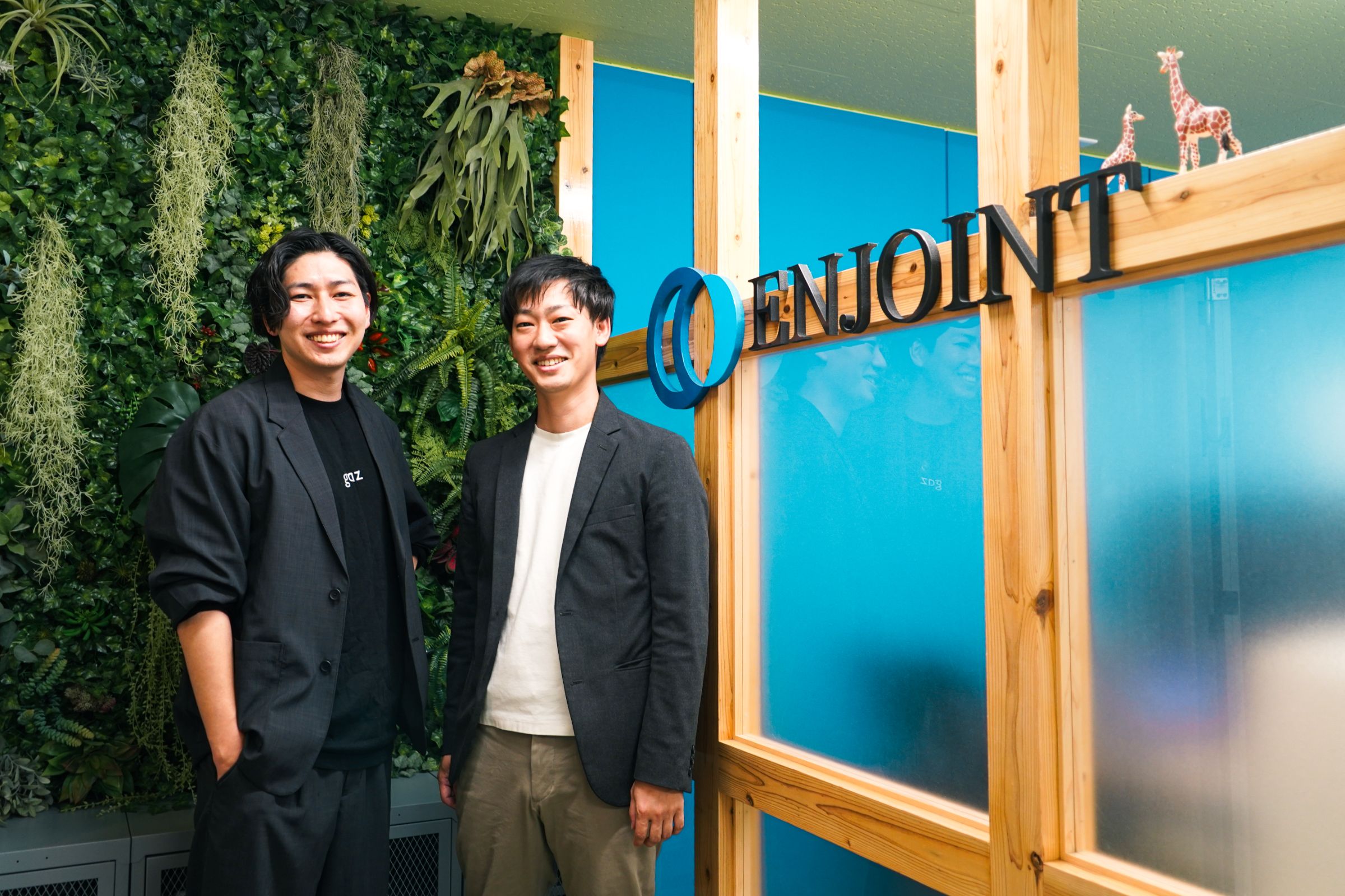 オフィスに掲出されているエンジョイントのロゴマークの前に立つ智原さんと吉岡さん。写真
