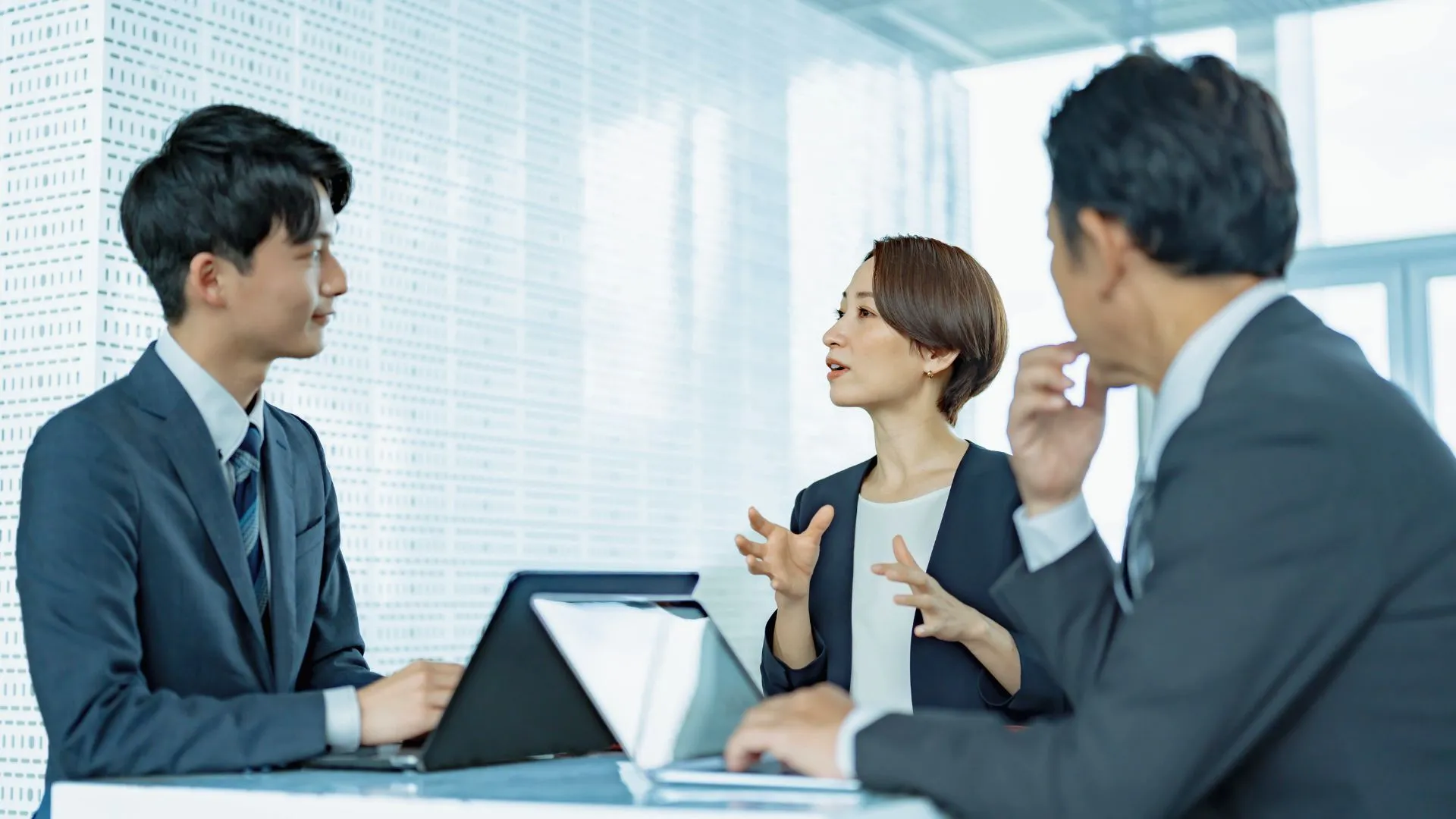 会議テーブルで女性が論点を整理して話し、同僚がノートPCを前に聞いている様子