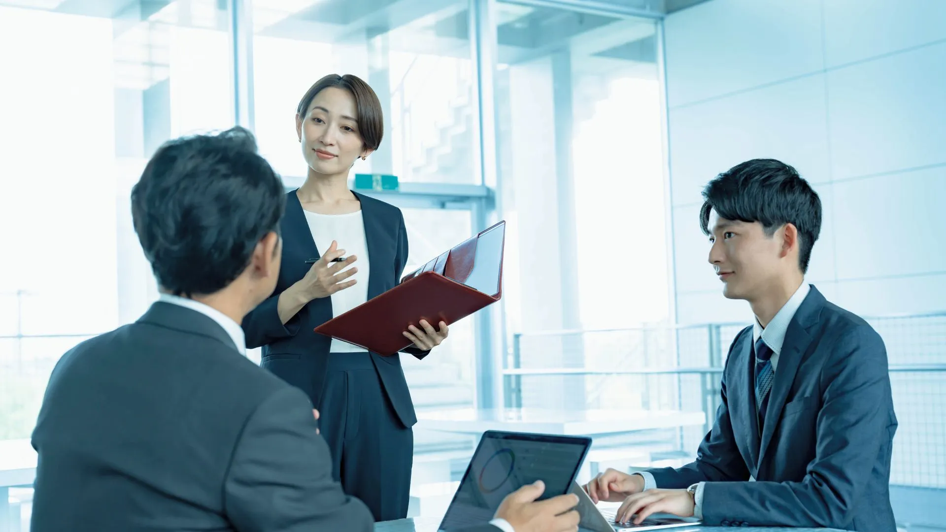資料ファイルを手にした女性が会議で要点を説明し、参加者が聞いている様子