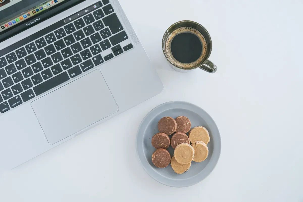 ノートパソコンのそばにコーヒーカップ・クッキーが盛られたお皿が並ぶ様子