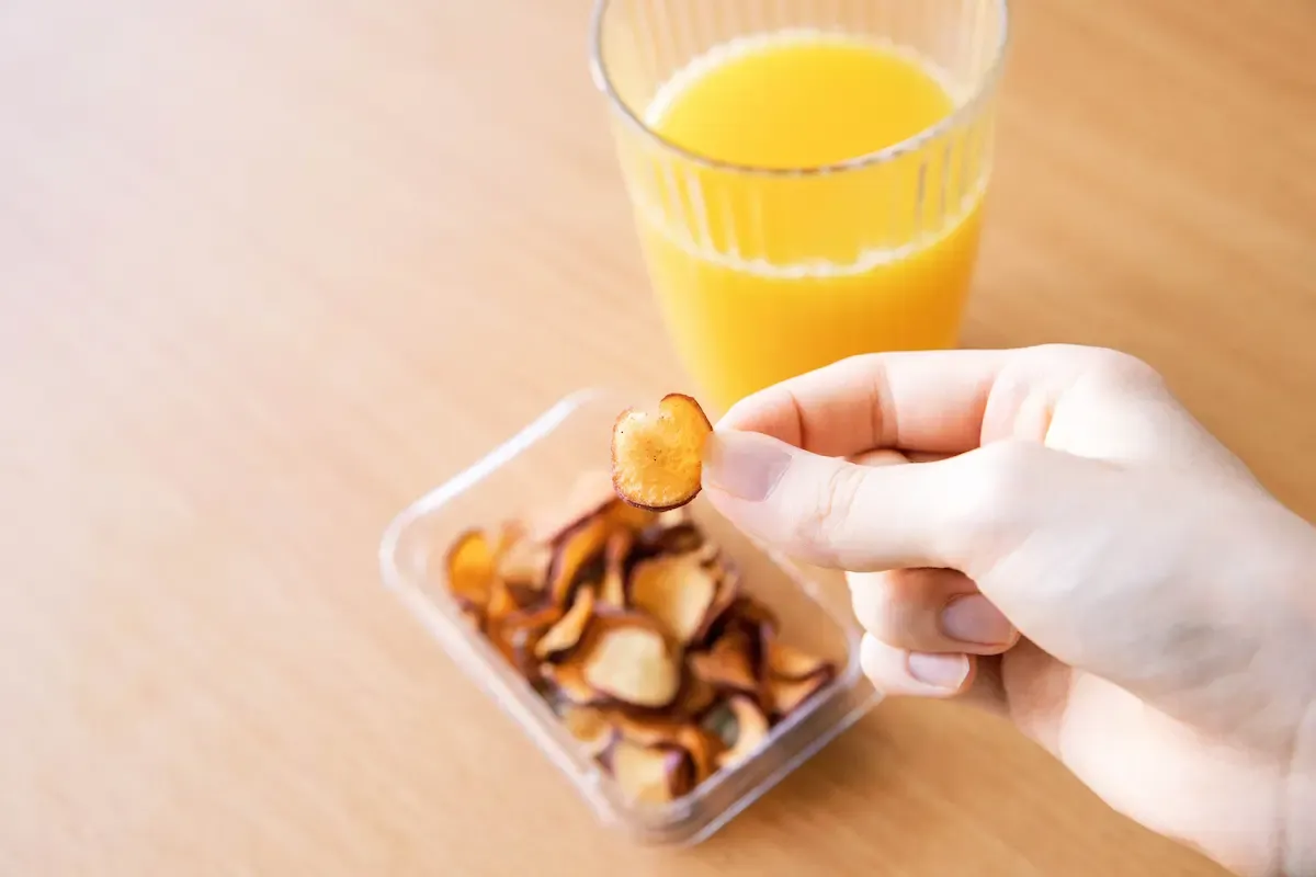 オレンジジュースのグラスのそばで、スナックミーのさつまいもチップスをつまむ様子
