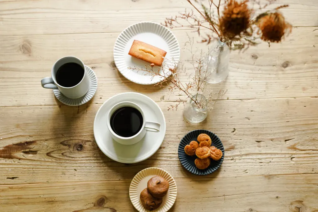ブラックコーヒーと焼き菓子、花瓶に生けた花が木製テーブルの上に並べられている様子。焼き菓子はフィナンシェ、パイ、クッキーの3種類。