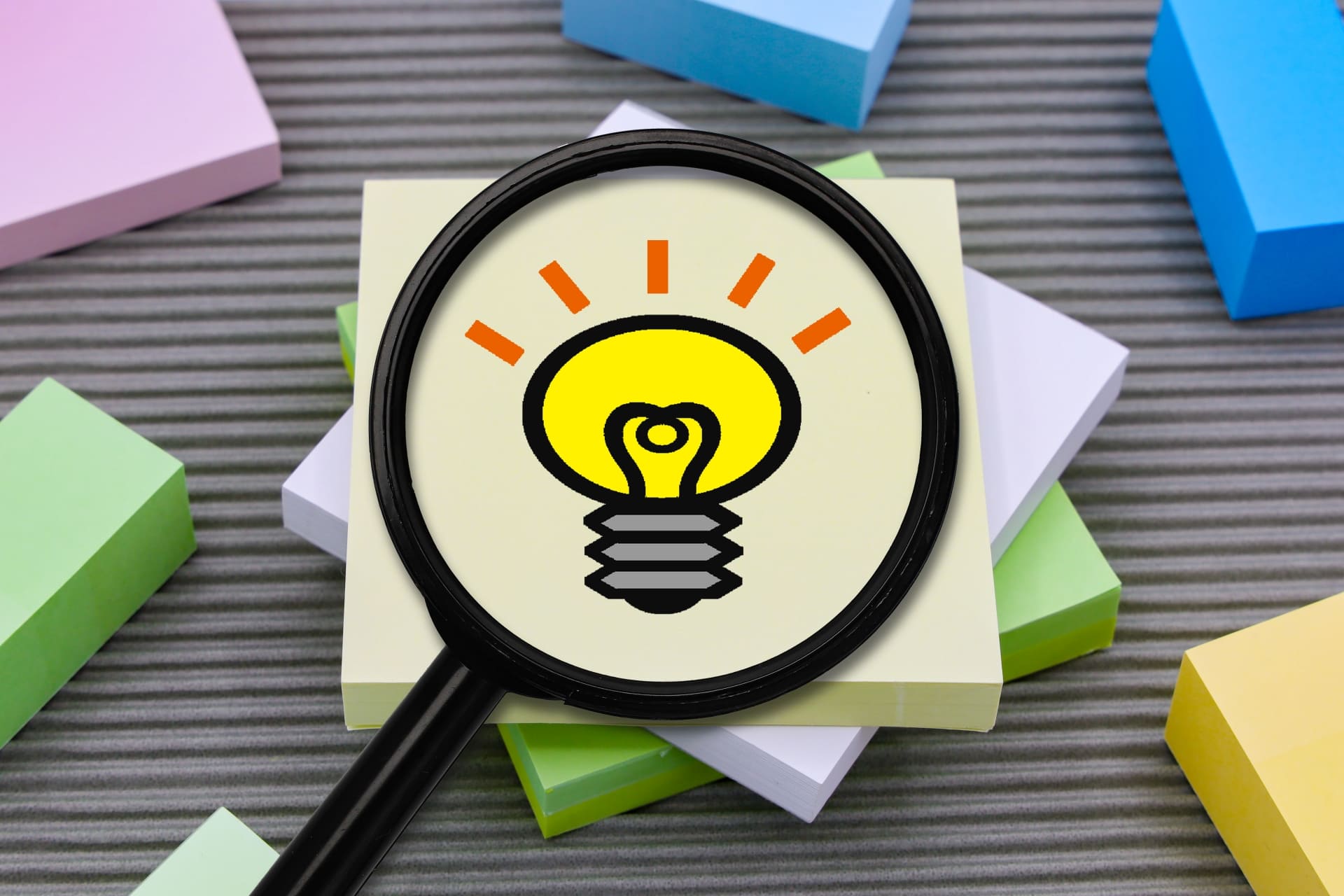 積み重なった正方形の付箋の上に書かれた電球マークにフォーカスをあてる虫眼鏡の画像