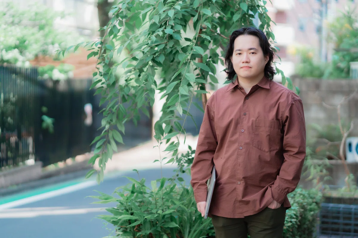 写真。オフィス外に出て、立ち姿で写真に映るFumitoさん。右手にはPCを抱えている。