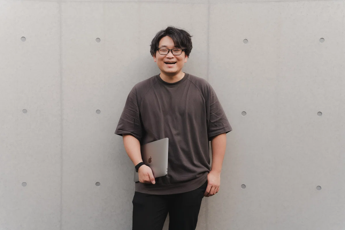 写真。打ちっぱなしコンクリート壁の前に立ち、笑顔を見せるMurakamiさん。右手にはPCを携えている。