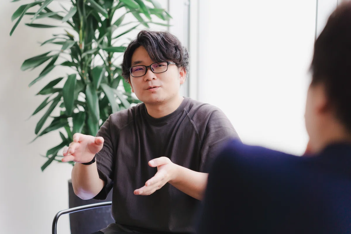 写真。インタビュイーに説明をするMurakamiさん。より頭を上げ、熱を込めて話している。