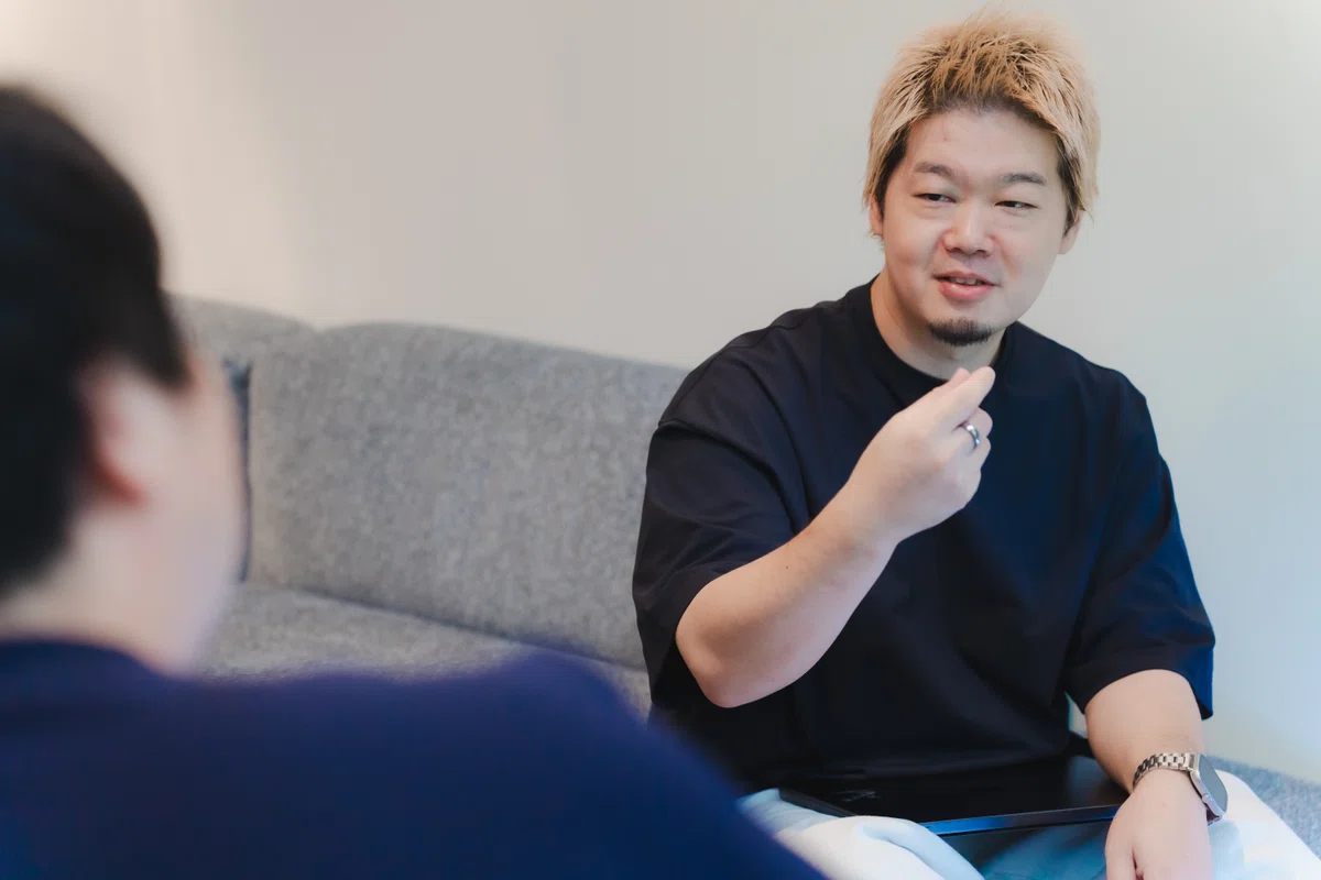 写真。目線をインタビュイーに向けて、身振り手振りで話をするYagiさん。