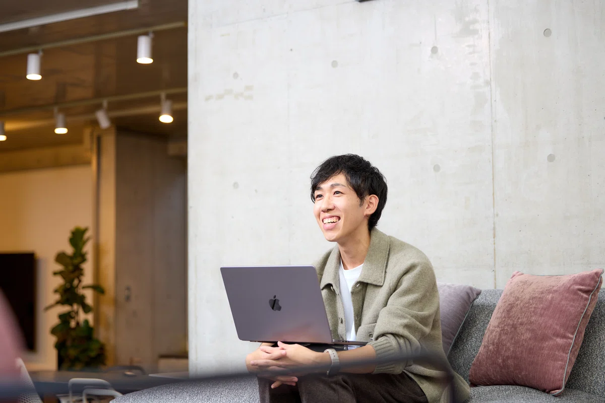 写真。PCを開き、笑顔でインタビューに答えるkamiyamaさん。前屈みでソファに腰掛けている。