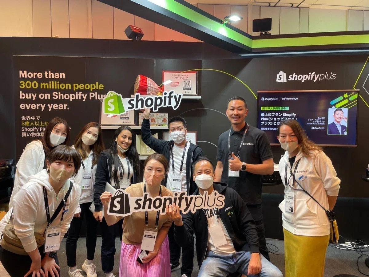 写真。前職「Shopify Japan」時代、展示会ブースで集合写真に写っているのEmaさん。中央左側、左から二人目に立ち、製品ロゴのボードを掲げている。