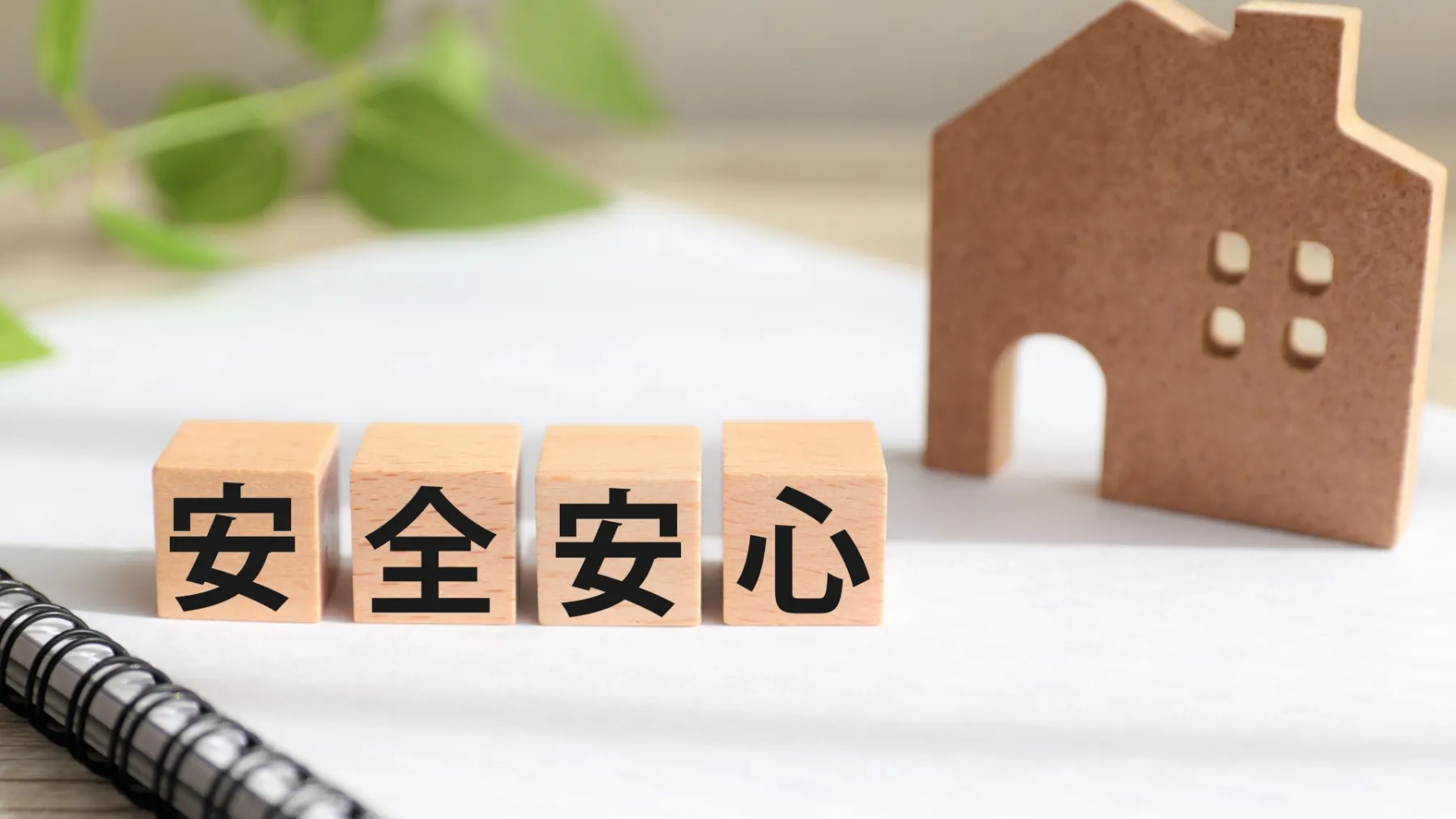 木の模型に安全安心と書かれた置物の奥に家のイメージ