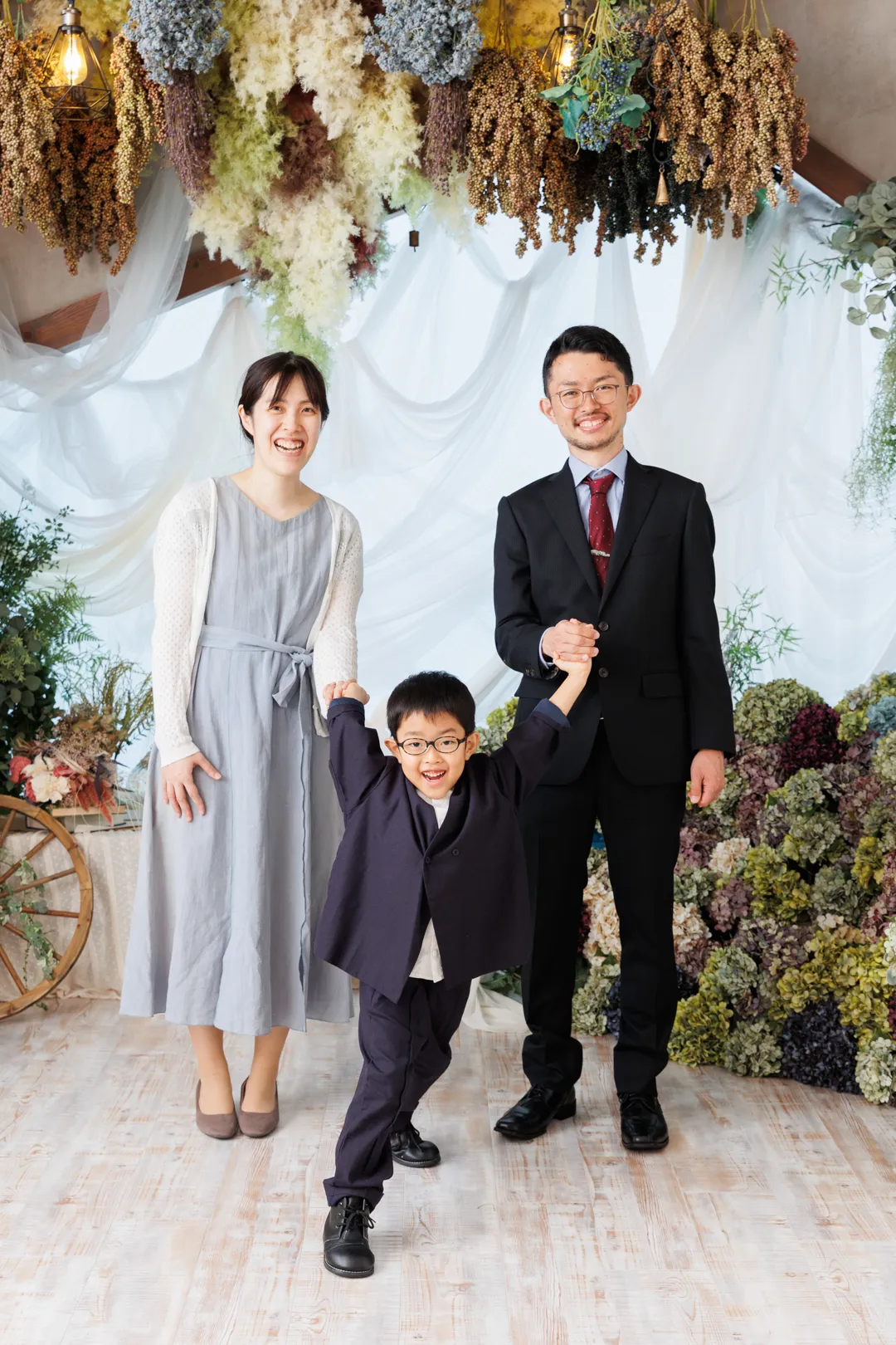 ドライフラワーの装飾を背景に両親と手をつなぐ紺色スーツ姿の男の子の七五三家族フォト（野々市市）｜ヤマダフォト