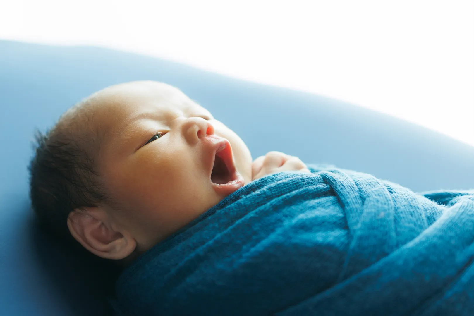 青い布団で大きくあくびをするブルーのおくるみに包まれた新生児(川北町)|ヤマダフォト