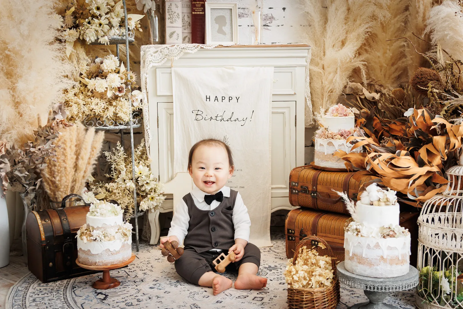 パンパスグラスとリースの飾りを背景にケーキに囲まれて座る蝶ネクタイ姿の男の子のバースデーフォト（小松市）｜ヤマダフォト