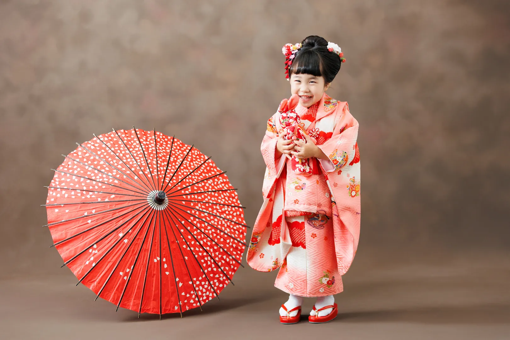 ピンクの花柄着物を着て大きな赤い和傘のそばに立つ3歳女の子の七五三記念写真（小松市）｜ヤマダフォト