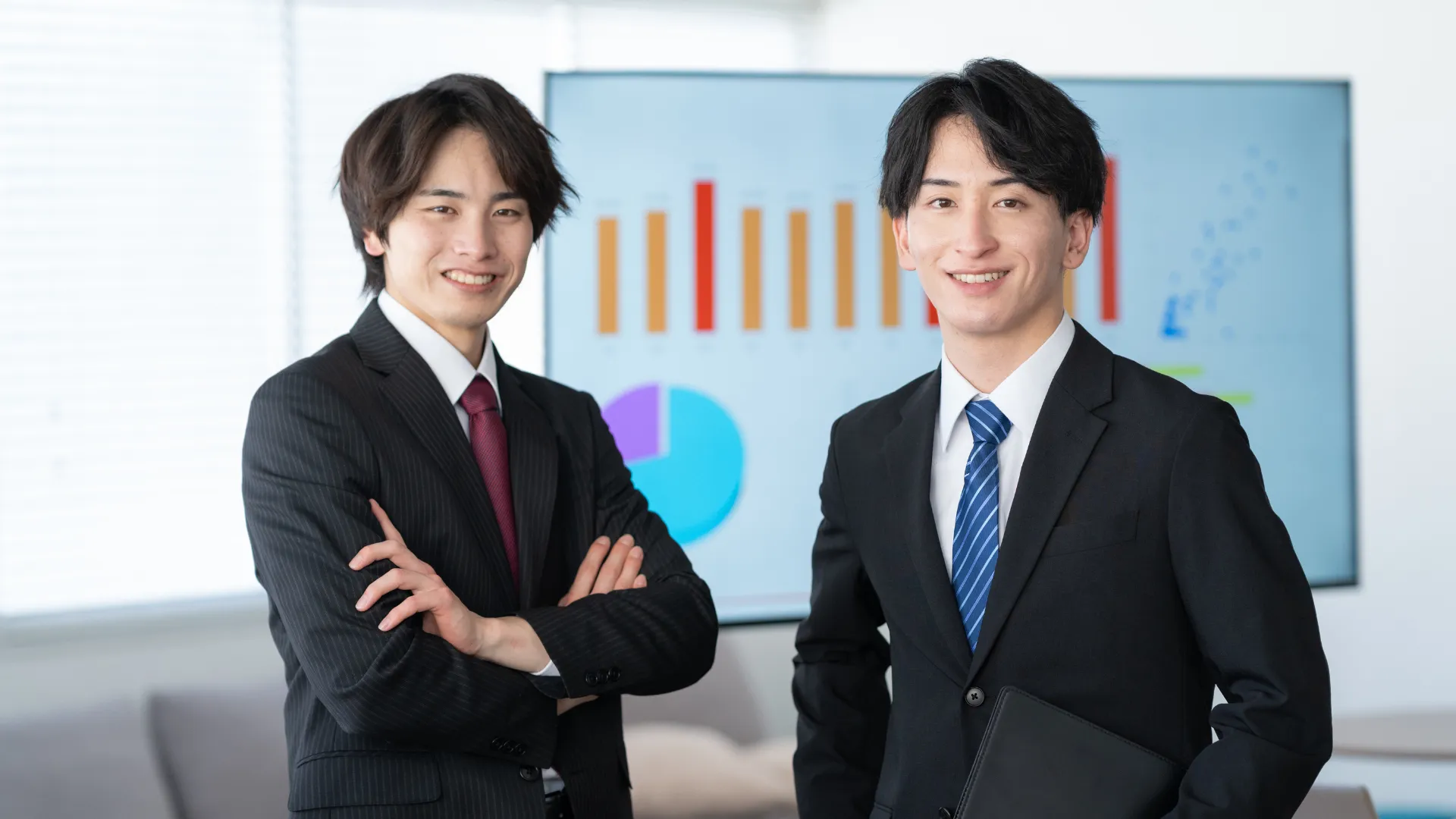グラフが表示されたモニターの前に立つ二人のビジネスパーソン。提案書の書き方を検討しているイメージ。