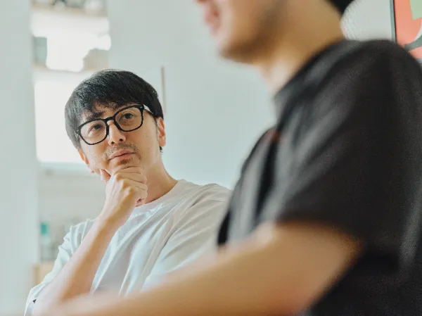 ALTはじまり: 眼鏡の奥から真剣な眼差しで大塚さんの話を聞きながら、顎に手を当てて考え込む犬飼さんの表情。ALTおわり