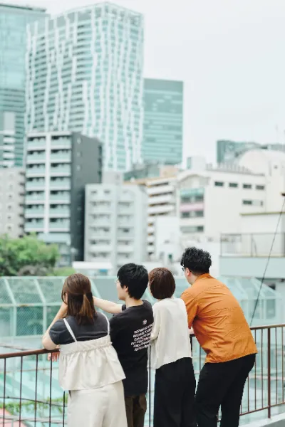 ALTはじまり: 屋上で街を見渡す4人の後ろ姿。右端に立つオレンジ色のシャツの人物が葉栗さん。ALTおわり