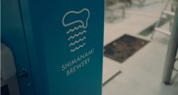 ALTはじまり：しまなみブルワリーの醸造所入り口にある、しまなみブルワリーのロゴ。瀬戸内海の雰囲気を感じる爽やかなブルーカラーの機械に、白い色でロゴが印字されている。ALTおわり