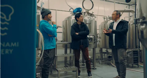 ALTはじまり：しまなみブルワリーのヘッドブルワー、松岡さんとアイティプラスの二人が、醸造所内で並んで笑顔で話している様子。無機質な機械が並ぶ醸造所のなか、松岡さんが着用するパーカーがしまなみブルワリーのブランドカラーである爽やかなブルーカラーで、とくに際立っている。ALTおわり