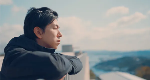 ALTはじまり：溝口さんが右を向いている横顔の写真。黒いダウンジャケットを着て、展望台の柵に腕を乗せながら尾道の街並みを眺めている。ALTおわり