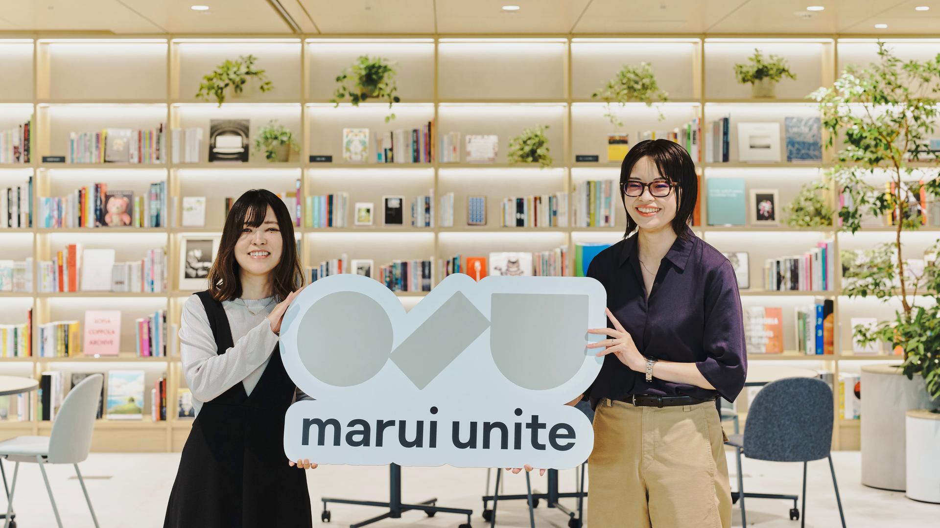 ALTはじまり:導入事例インタビューサムネイル画像。株式会社マルイユナイトの風間さん(右)と、株式会社Mutureの新井さん(左)の二人が、「marui unite」のロゴボードを持って、本棚のあるラウンジに立っている。ALTおわり