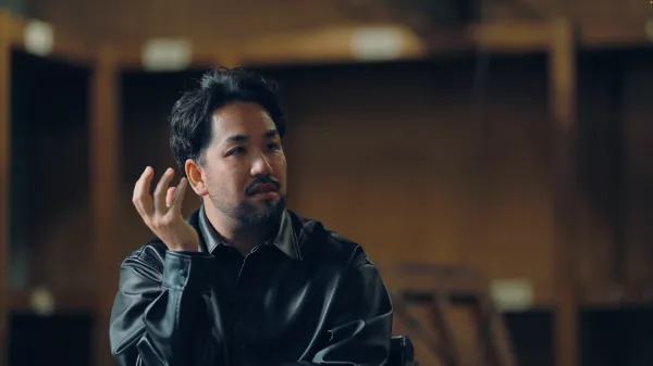 ALTはじまり：Studioの代表、石井が話している様子。右手を顔の横に置き、考えながら話している。黒いレザージャケットを着ている。ALTおわり