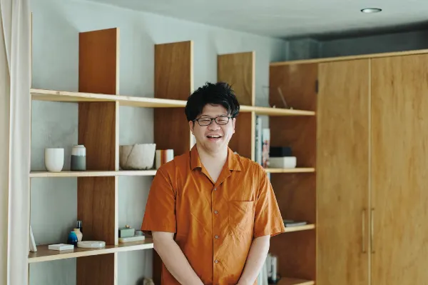 ALTはじまり: 棚の前で笑顔を見せる葉栗さん。カジュアルな雰囲気の室内で撮影。ALTおわり